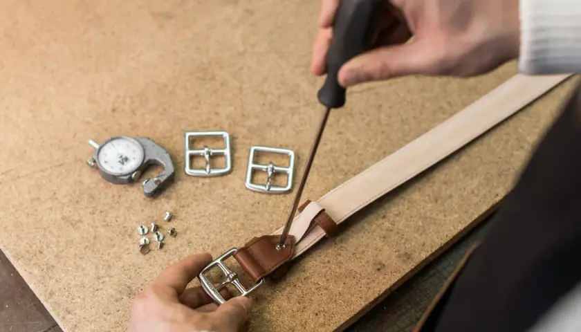 Artisan travaillant sur une ceinture en cuir, utilisant un tournevis pour fixer une boucle. Outils de mesure et boucles supplémentaires à proximité.