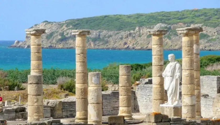 Ruinas romanas en el área de Bolonia, con columnas de piedra y una estatua blanca frente al mar azul y una colina verde.
