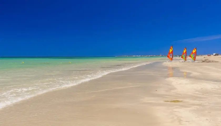 Piękna plaża w Tunezji w czerwcu. Trzy kolorowe deski windsurfingowe stoją na piasku, a turkusowa woda łagodnie faluje.