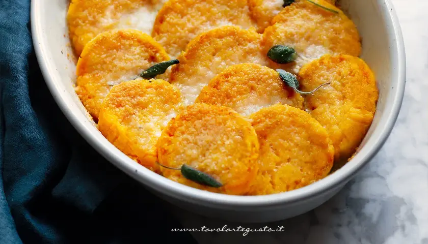 Gnocchi di zucca dorati e gratinati, guarniti con foglie di salvia, in una pirofila bianca.
