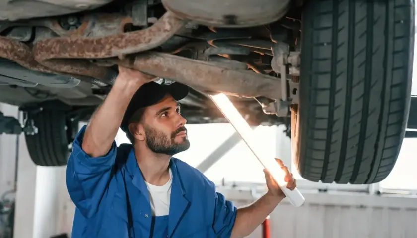 Mechanik w niebieskim kombinezonie oświetla latarką podwozie samochodu, sprawdzając jego stan. Dowiaduje się, ile trwa przegląd samochodu.