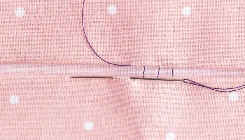 Close-up of a needle and purple thread creating an invisible hem stitch on pink polka-dot fabric.