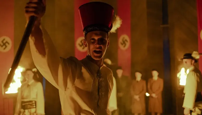 Młody mężczyzna w czerwonym nakryciu głowy z piórem wznosi płonący kij. W tle widać flagi ze swastykami, sugerujące scenę z filmu "Biała odwaga".