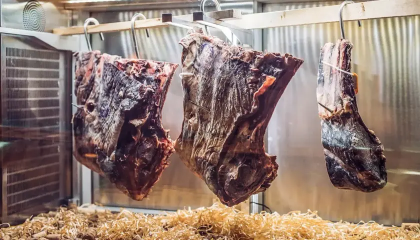 Drei große Fleischstücke hängen in einer Kühlkammer, bereit zum **Fleisch fermentieren**. Darunter liegen Holzspäne und Korken.