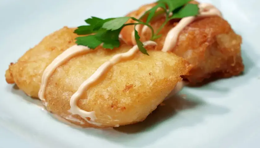 Dos trozos de bacalao rebozado, dorados y crujientes, servidos con una salsa cremosa y perejil fresco.