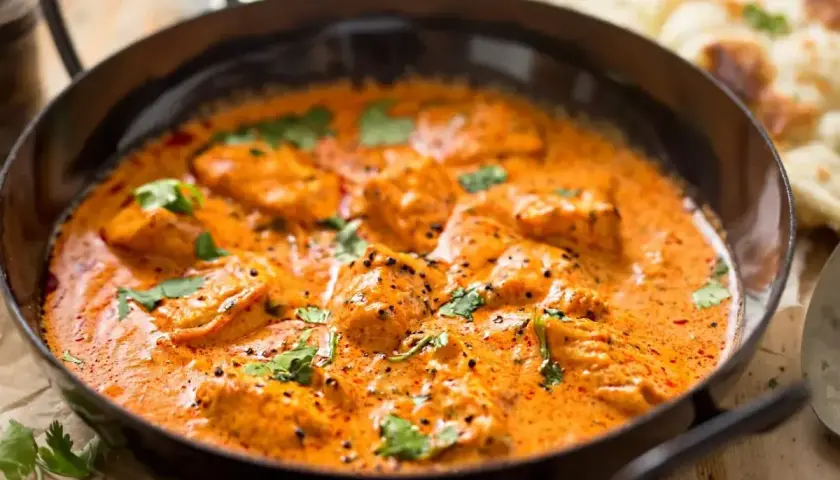 Aksamitny kurczak w sosie butter masala, ozdobiony świeżą kolendrą, podany w czarnej patelni. Obok widać kawałki chleba naan.