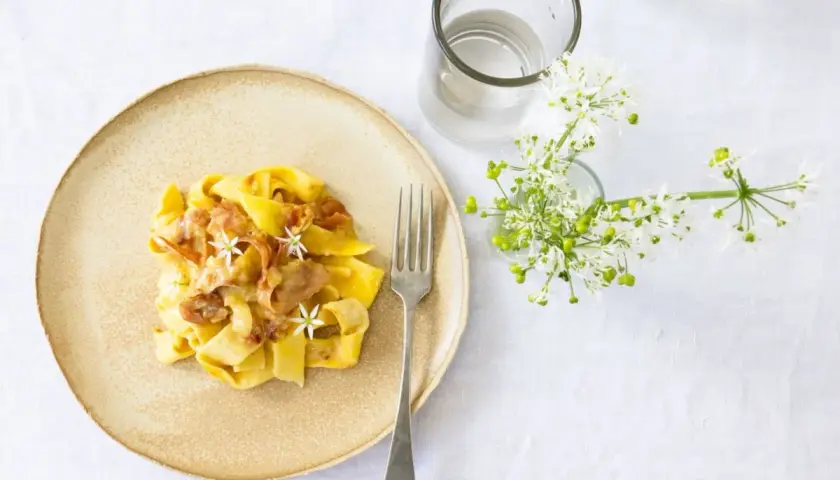 Ein Teller mit cremiger Pasta, die wie Fettuccine aussieht, garniert mit zarten Blüten und Speck. Daneben ein Glas Wasser und ein Strauß weißer Blumen.