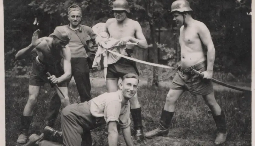 Fünf Männer, einige mit Helmen, posieren mit Waffen. Ein Mann hält eine Puppe. Die Szene erinnert an die Verbrechen der Wehrmacht.