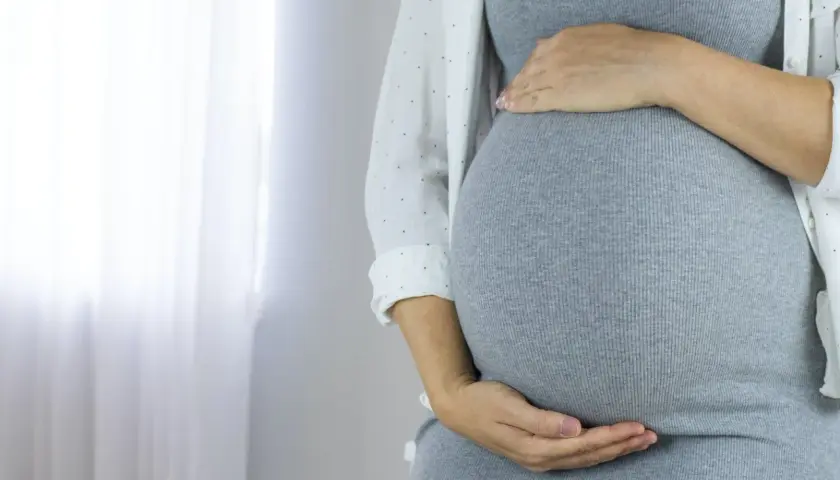 Zbliżenie na brzuch ciężarnej kobiety w szarej sukience, trzymający się za brzuch.