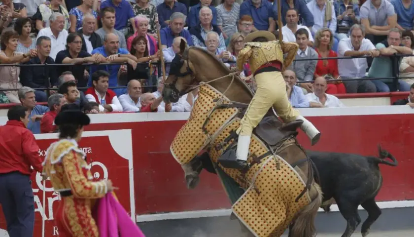 Un rejoneador, quizás Ana Romero, monta un caballo cubierto con un capote mientras un toro se acerca.