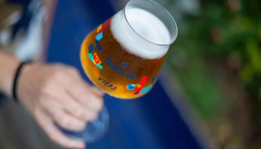 Main d'une personne tenant un verre de bière Gallia, orné de motifs colorés. La bière est dorée avec une mousse blanche.