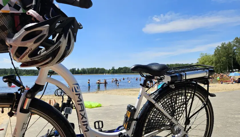 Rower z kaskiem czeka na plaży nad jeziorem, gdzie w upalny dzień kąpie się wiele osób. Idealne miejsce na wycieczkę rowerową i relaks w Puszczy Dulowskiej.