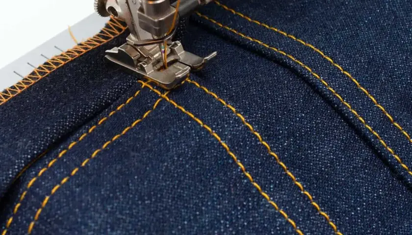 A sewing machine's topstitching foot creates neat, parallel yellow stitches on dark denim fabric, adding a professional finish.