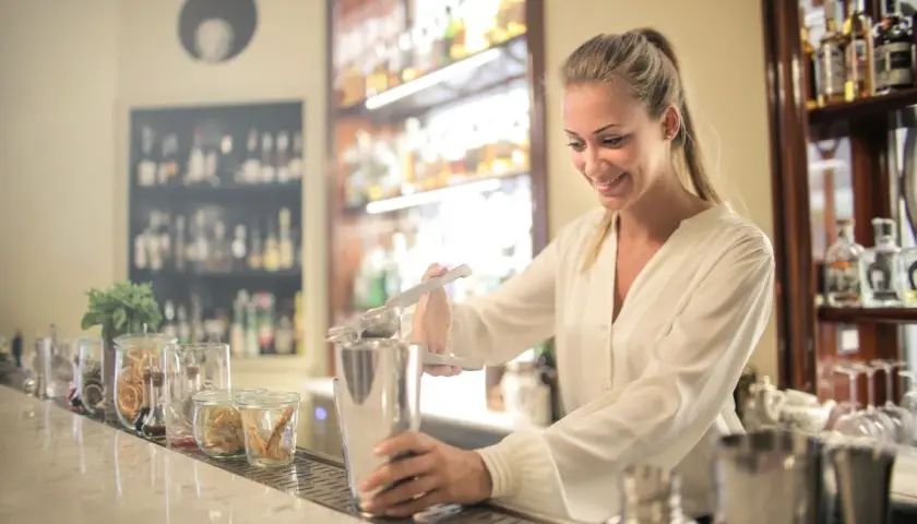 Uśmiechnięta barmanka przygotowuje drinka, ucząc się, jak zostać barmanem. Za nią półki z alkoholem.