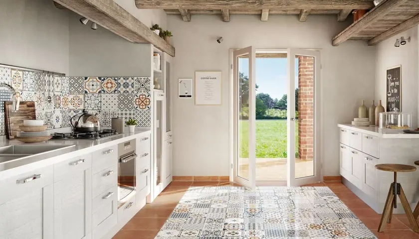 Cucina rustica con pavimento decorato, pensili bianchi e un'ampia porta finestra che si apre su un prato verde. La posa battiscopa è curata nei dettagli.