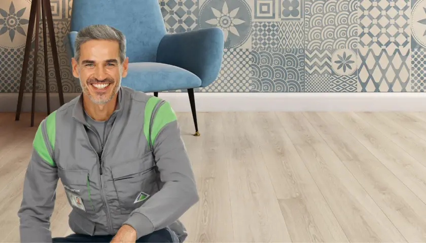 Un uomo sorridente in gilet da lavoro, pronto per il montaggio SPC. Sfondo con poltrona blu e parete decorata.