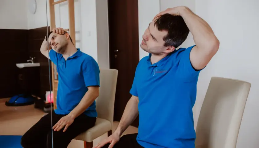 Mężczyzna w niebieskiej koszulce wykonuje ćwiczenia na kręgosłup szyjny, delikatnie przechylając głowę do boku.