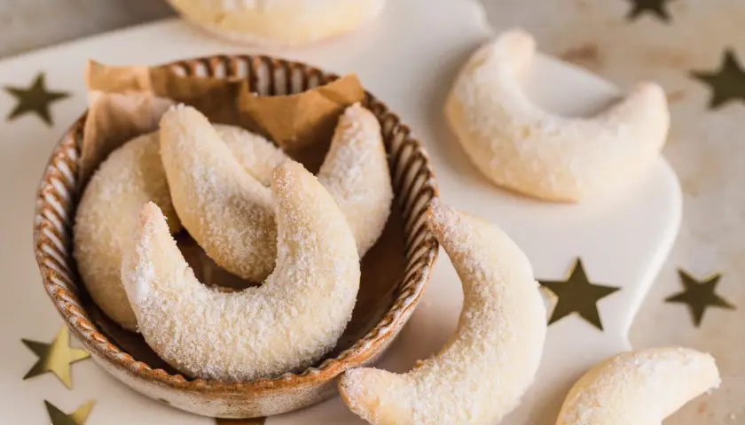 Vegane Vanillekipferl rezept: zarte, mondförmige Kekse, bestäubt mit Puderzucker, liegen in einer Schale und auf einem Brett, umgeben von goldenen Sternen.