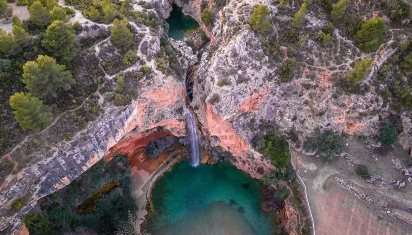 Cascada cayendo en una poza turquesa, rodeada de rocas rojizas y vegetación. Un rincón secreto, como la cueva turche, esperando ser descubierto.