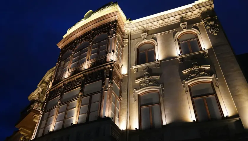Elegancka kamienica nocą, oświetlona ciepłym blaskiem lamp miejskich. Widać bogate zdobienia fasady i duże, łukowe okna.