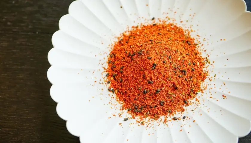 A pile of vibrant Japanese 7 spice blend, featuring chili flakes and black sesame seeds, sits on a white, fluted dish.