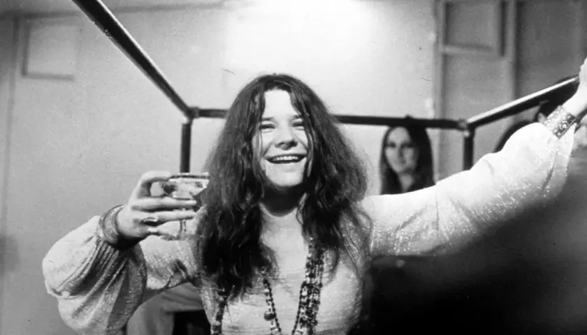 Janis Joplin, z kieliszkiem w dłoni, uśmiecha się szeroko. Jej charakterystyczne włosy opadają na ramiona, a na szyi wisi naszyjnik. W tle widać inną kobietę. To zdjęcie przypomina o jej niezapomnianych **janis joplin utworach**.