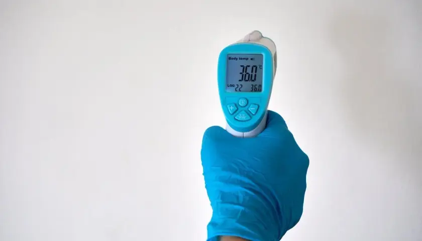 Ręka w niebieskiej rękawiczce trzyma termometr bezdotykowy, pokazujący 36.0°C. Idealne miejsce, gdzie mierzyć temperaturę na czole lub skroni.