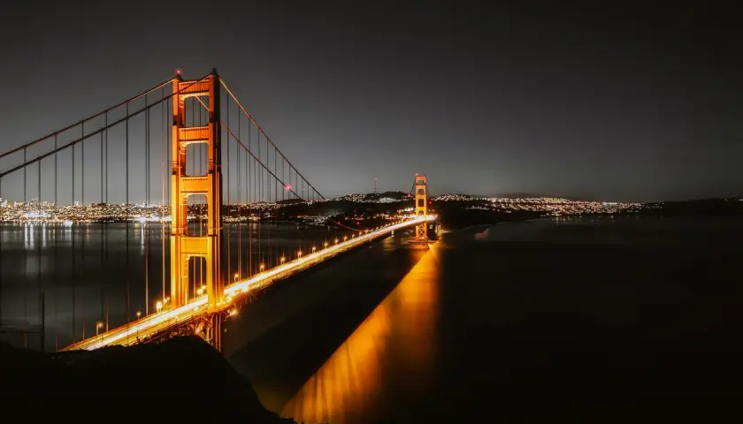 Złota Brama nocą, oświetlona, nad wodą. W oddali widać miasto w Kalifornii, San Francisco, z migoczącymi światłami.