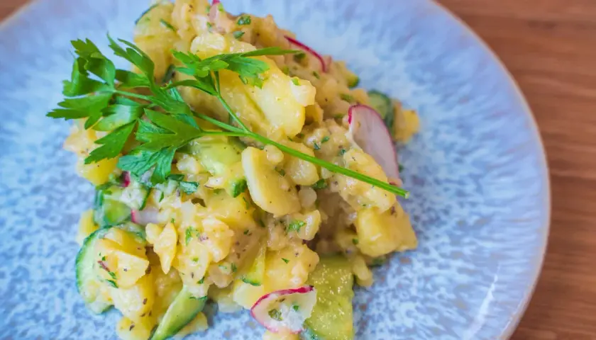 Ein köstlicher Kartoffelsalat mit Gurken, Radieschen und frischer Petersilie auf einem blauen Teller.