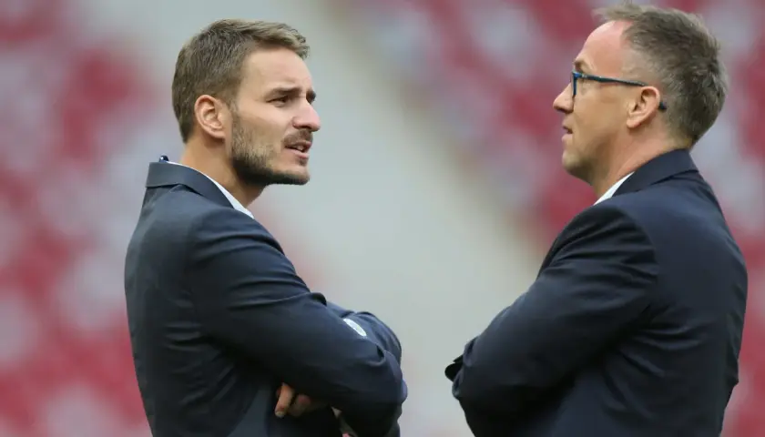 Dwaj mężczyźni w garniturach rozmawiają na tle stadionu. Jeden z nich, z zarostem, to dyrektor sportowy.