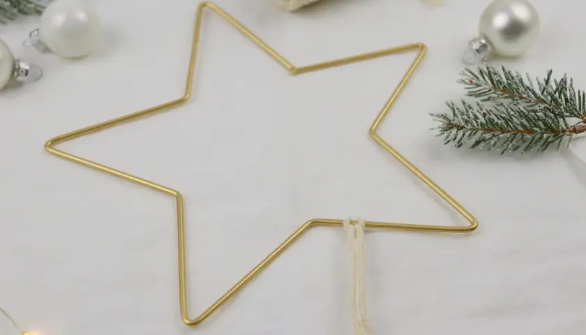 Goldener Sternrahmen für Makramee Weihnachten, umgeben von Tannenzweigen und weißen Kugeln.