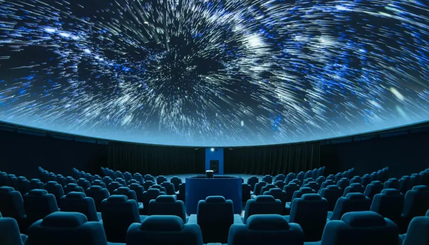 Wnętrze planetarium, gdzie na kopule wyświetlane są rozgwieżdżone niebo i galaktyki. Widownia z niebieskimi fotelami czeka na seans.