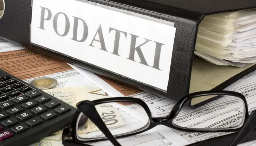 Segregator z napisem "PODATKI", kalkulator, okulary i dokumenty. Obok leży moneta, symbolizująca potencjalną kwotę zmniejszającą podatek.