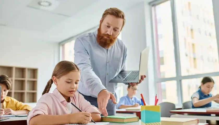 Nauczyciel z brodą, trzymający laptopa, pomaga uczennicy w pisaniu. W tle inni uczniowie. To jego legitymacja nauczyciela.