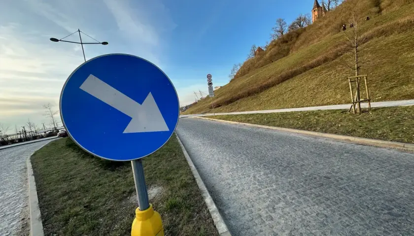Niebieski znak z białą strzałką w prawo wskazuje kierunek jazdy. Droga z kostki brukowej prowadzi pod górę, gdzie widać zarys kościoła. Taryfikator opłat za parkowanie jest gdzieś w pobliżu.