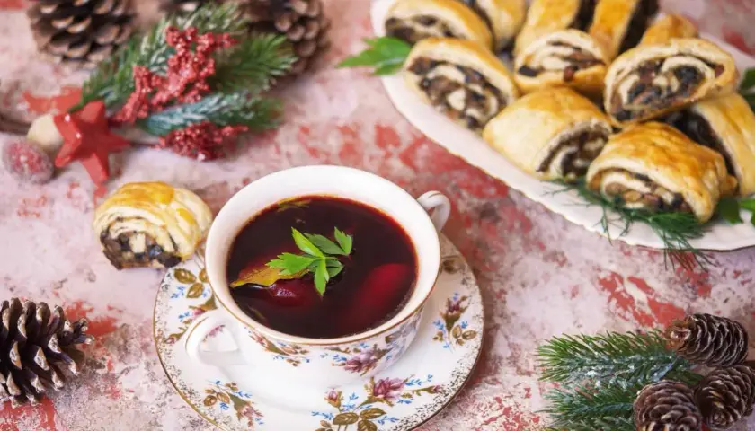 Aromaticzny barszczyk czerwony z pasztecikami, ozdobiony gałązką zieleni, czeka na świątecznym stole wśród szyszek i gałązek.