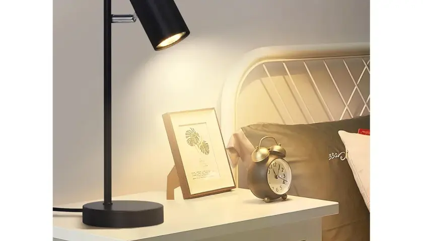 Lampara mesilla negra ilumina un cuadro y un despertador vintage en una mesita de noche junto a una cama.