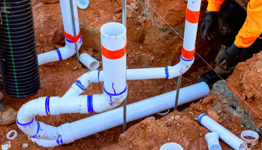 Prace hydrauliczne w toku: łączenie rur pcv w wykopie. Widać fragmenty instalacji kanalizacyjnej i drenażowej.