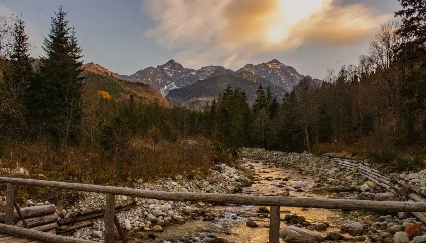 Jesienne Tatry w październiku. Drewniany mostek nad kamienistym potokiem, w tle góry ze śniegiem i złote chmury.