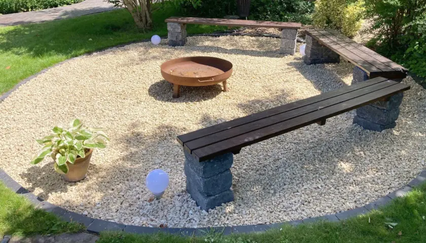 Gemütliche Kies Garten Sitzecke mit Feuerstelle und Holzbänken. Kugelleuchten sorgen für stimmungsvolles Licht.