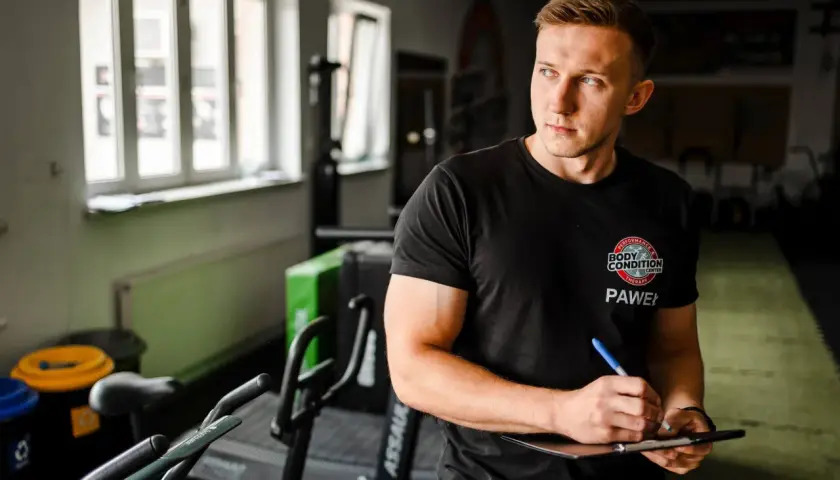 Trener Paweł tworzy spersonalizowany plan treningowy na biceps, notując ćwiczenia na tablicy.