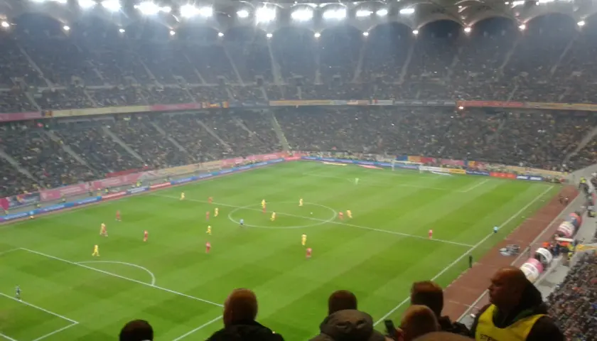 Mecz piłki nożnej na stadionie, tłumy kibiców, flagi Polski i Rumunii.