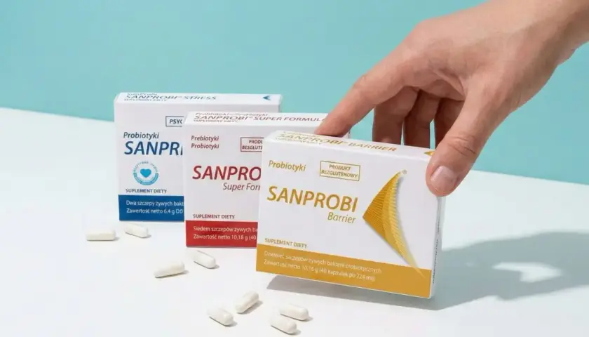 Dłoń sięga po probiotyk na zaparcia Sanprobi Barrier. Na stole leżą też opakowania Sanprobi Super Formula i Stress.