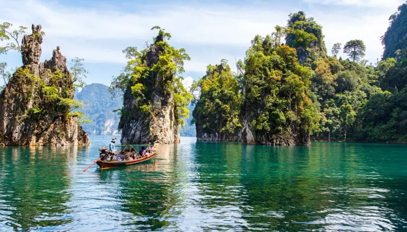 Bootfahrt durch smaragdgrünes Wasser im **Thailand Regenwald**, vorbei an steilen, bewachsenen Felsen unter blauem Himmel.