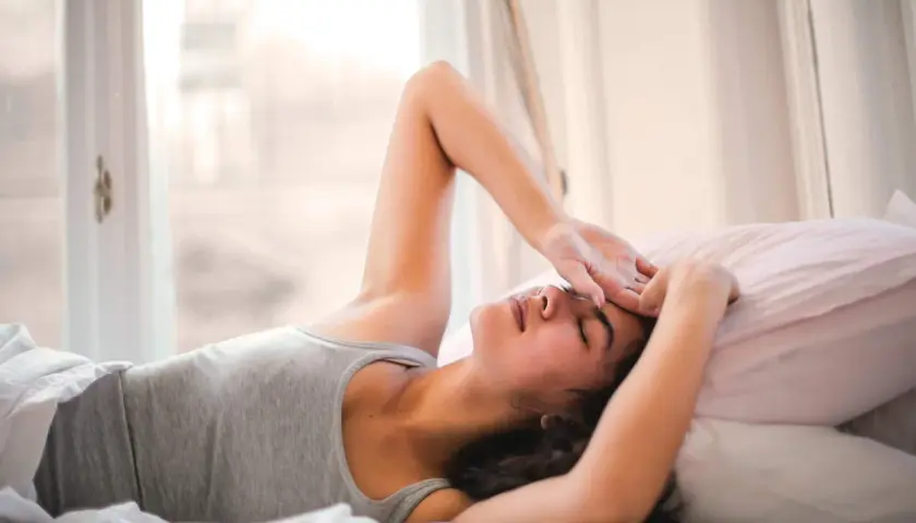 Ragazza sdraiata a letto, con la mano sulla fronte, sembra che non riesca a svegliarsi la mattina. Luce soffusa dalla finestra.