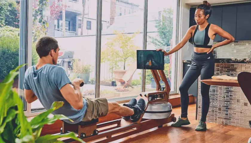 Para ćwiczy na ergometrze wioślarskim, który wygląda jak rower dla bliźniaków. Kobieta stoi obok, patrząc na ekran.
