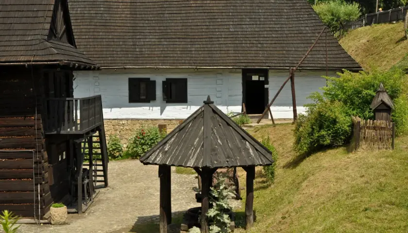 Skansen w Dobczycach: drewniane chaty, studnia i zielone wzgórza tworzą sielski krajobraz.