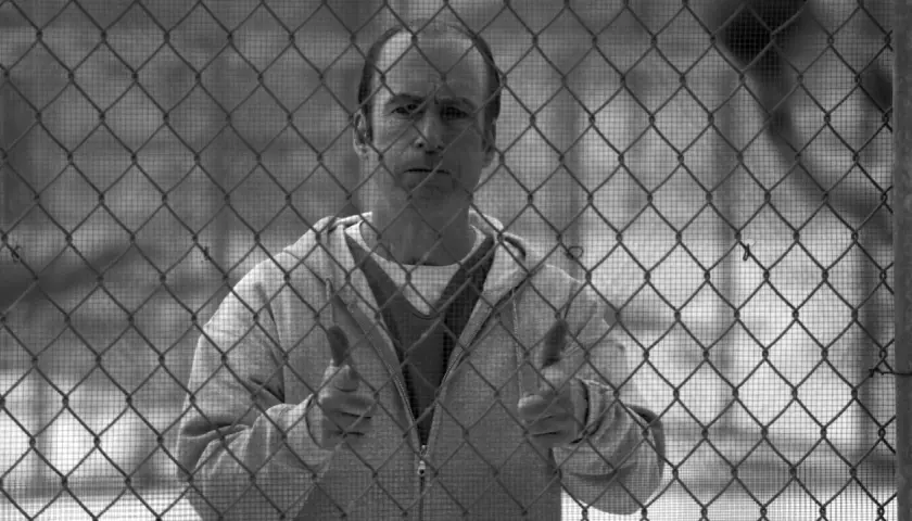 Un uomo dietro una rete metallica, con un'espressione pensierosa. Il suo gesto con i pollici in su suggerisce un "tutto bene" che contrasta con la sua situazione, evocando il **better call saul significato** di apparenza ingannevole.