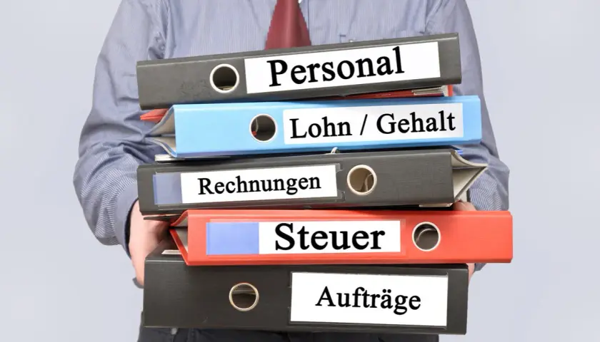 Ein Mann hält einen Stapel Ordner mit den Beschriftungen "Personal", "Lohn / Gehalt", "Rechnungen", "Steuer" und "Aufträge". Die Ordner stapeln sich und deuten auf die Komplexität der Verwaltung des zu versteuernden Einkommens hin.