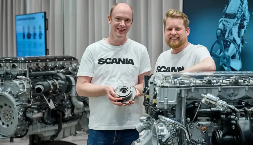 Dwóch mężczyzn w koszulkach Scania prezentuje części silnika. W tle widać potężny silnik Scania V8.